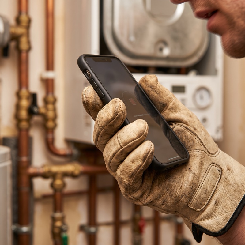 A gloved hand holds a phone up to speak into it, in front of a boiler and copper pipework.